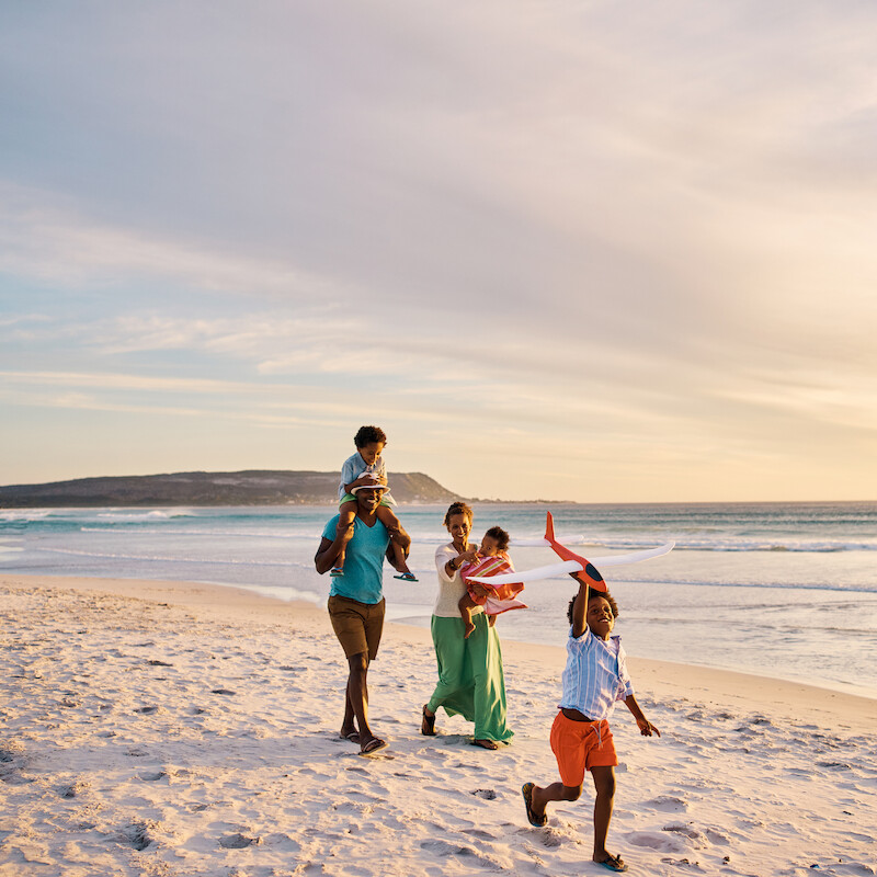 A family is enjoying a beach walk at sunset. One child is on an adult's shoulders, while another runs ahead with a hat.