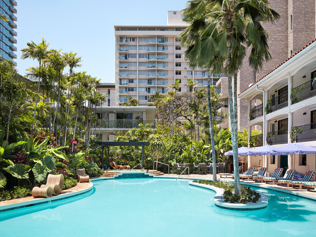 La imagen muestra el área de la piscina de un resort tropical con tumbonas, palmeras y edificios circundantes, creando una atmósfera serena y acogedora.