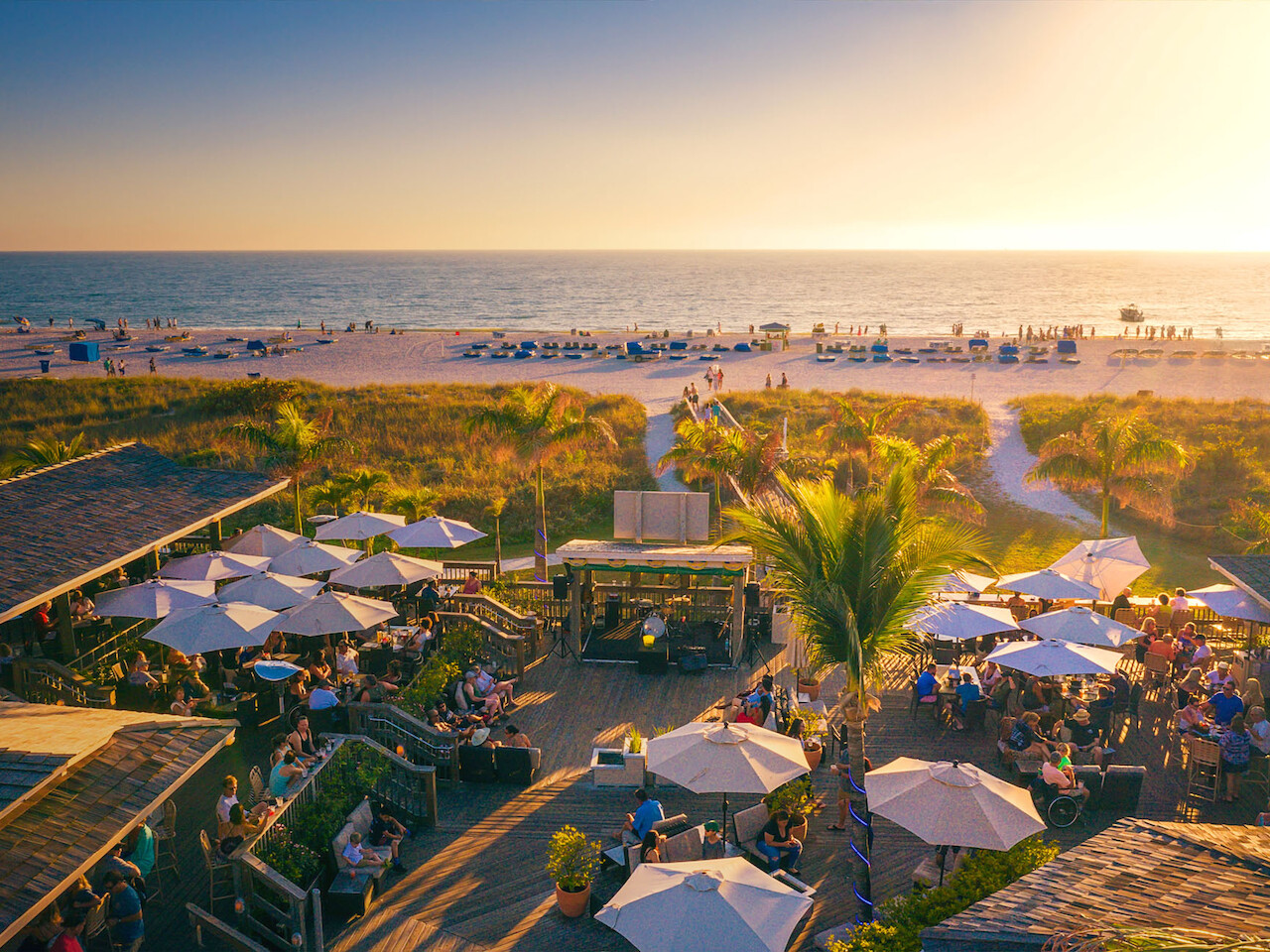 A beach scene with people dining under umbrellas, palm trees, and a beautiful sunset over the ocean, with distant lounge chairs on the sand.