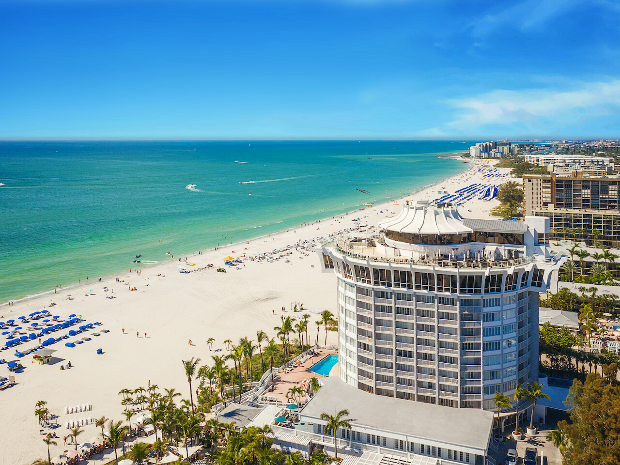 A beachfront scene with a circular hotel, palm trees, beach umbrellas, turquoise waters, and a clear blue sky ending the sentence.