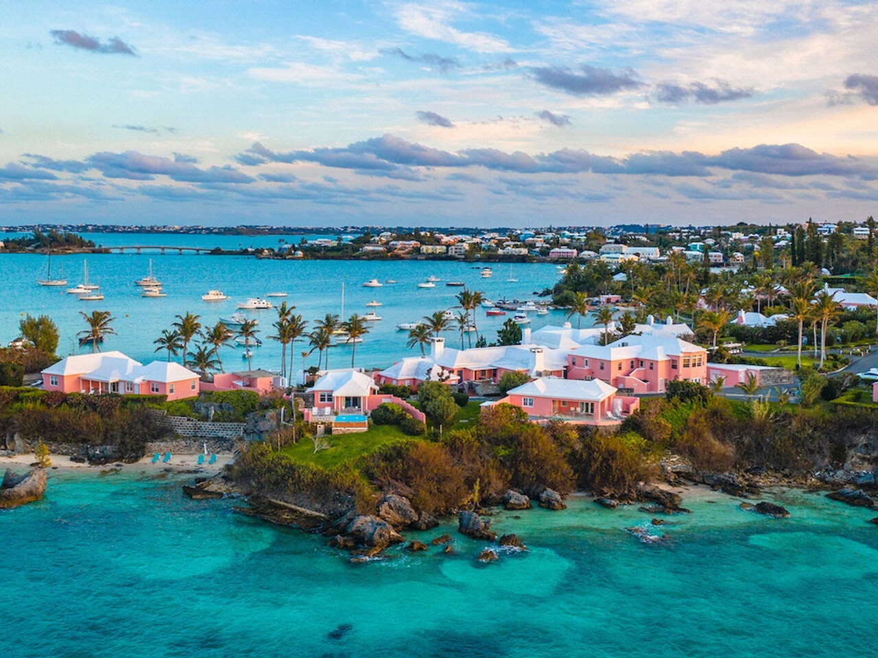 La imagen muestra una escena costera con casas coloridas, agua azul clara, barcos y exuberante vegetación con un cielo parcialmente nublado como telón de fondo.