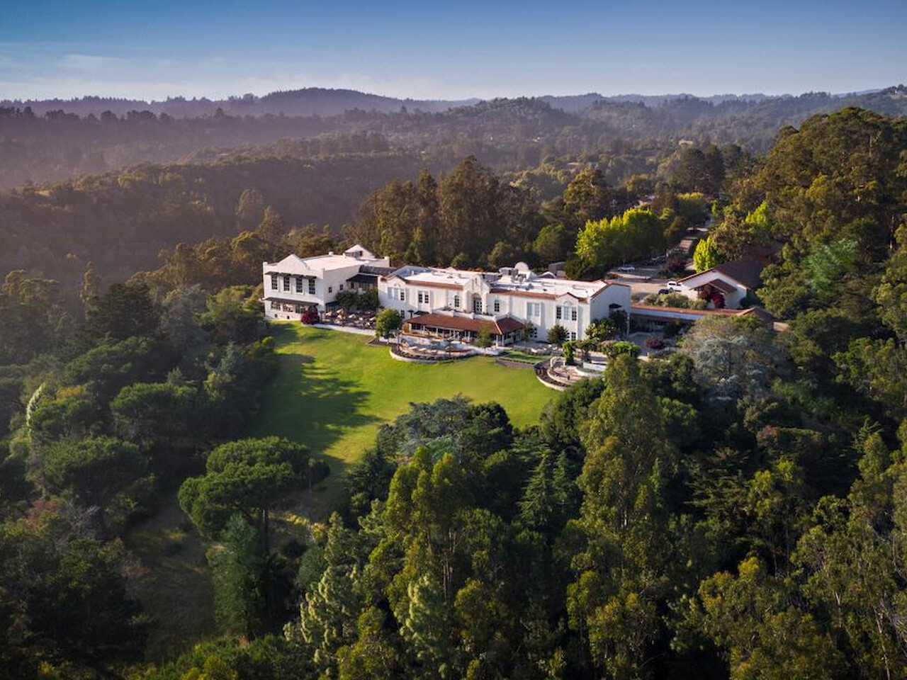 An aerial view of a large, luxurious mansion surrounded by lush greenery, with a scenic backdrop of forested hills and clear skies.