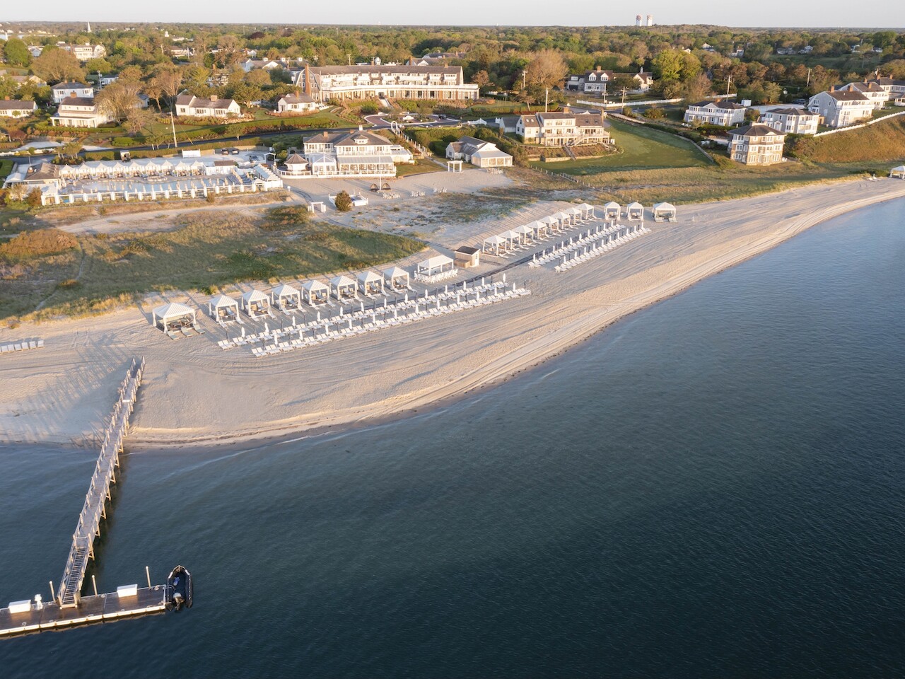 Aerial view of a beach with rows of cabanas, lounge chairs, a pier, and nearby shoreline houses. Serene setting next to the ocean.
