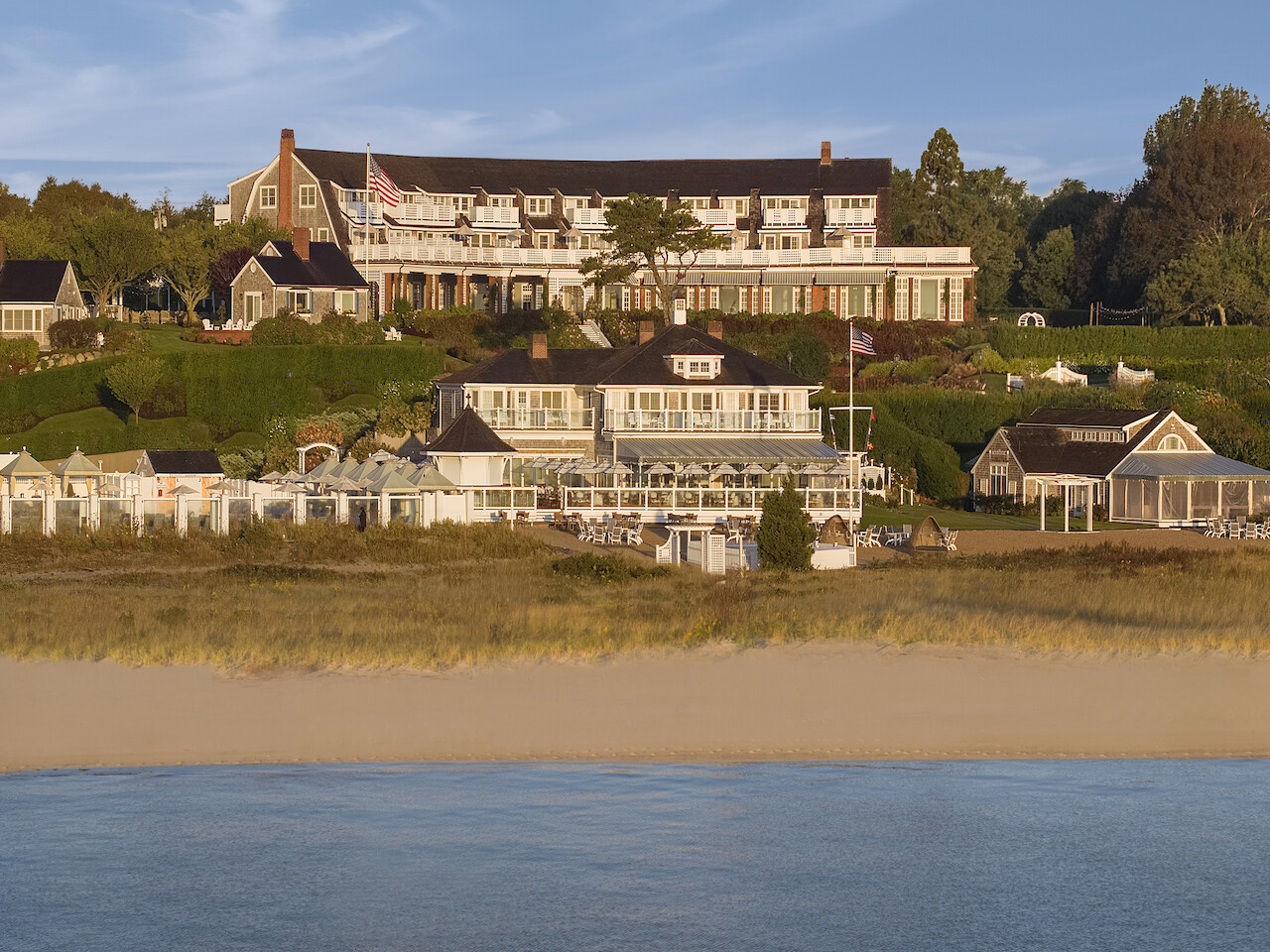 A large building situated on a hill overlooks a sandy beach and water, surrounded by greenery and smaller structures.