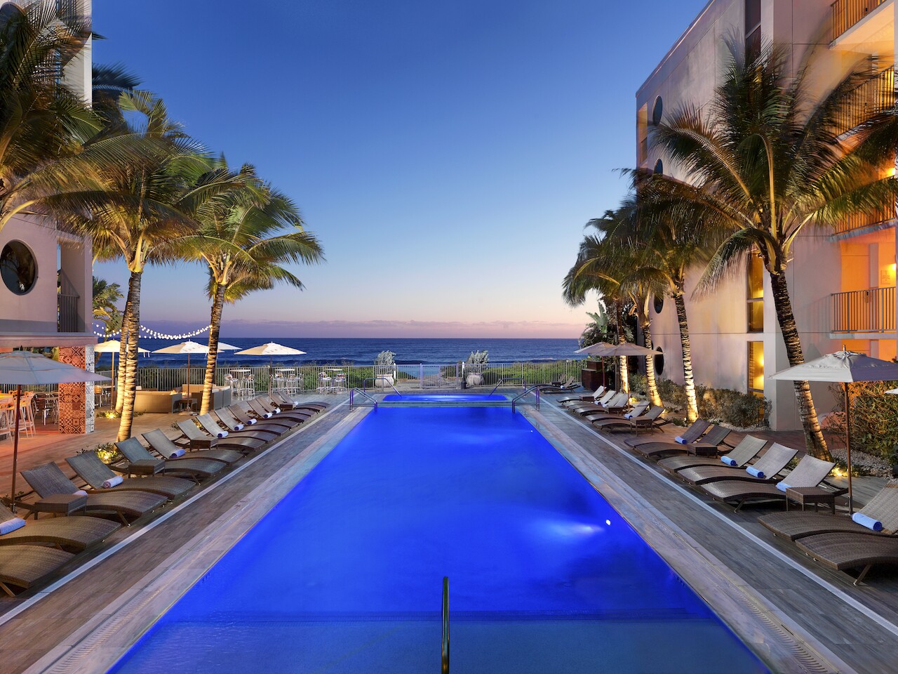 A luxurious outdoor pool surrounded by lounge chairs and palm trees, overlooking the ocean at sunset with sky hues.