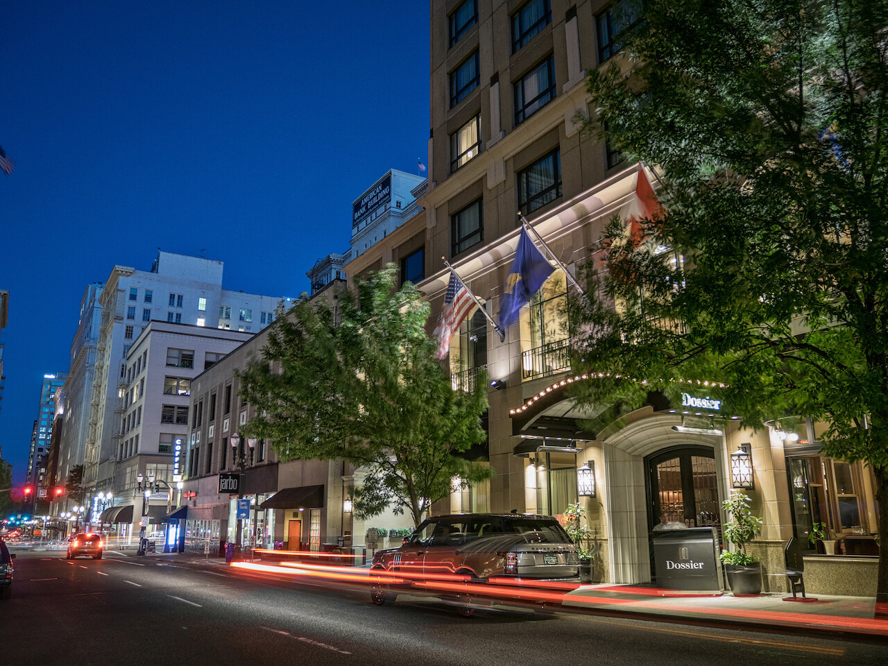 A nighttime city street with buildings, cars, and flags in front. One building&amp;#039;s sign reads &amp;quot;Theodore.&amp;quot; Trees and light trails from cars are visible.