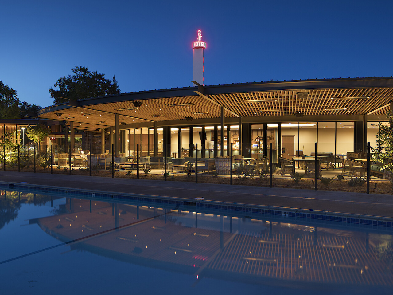 The image shows a modern building with large glass windows, an outdoor patio area, and a pool in the foreground, captured at dusk with lights on.