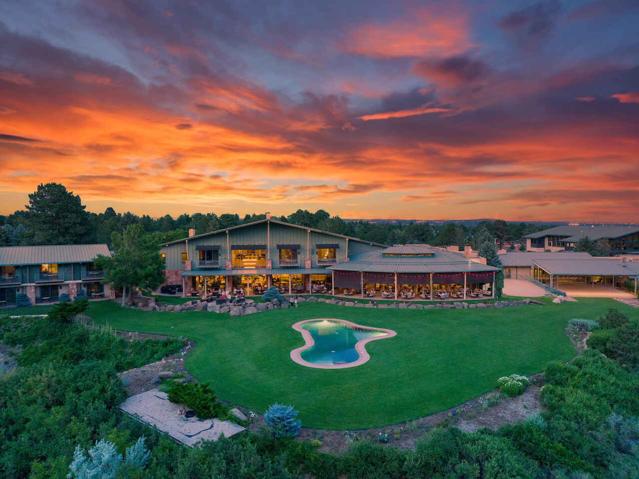 A luxurious house with large windows, a lush green lawn, a uniquely-shaped pool, and a scenic sunset in the background.