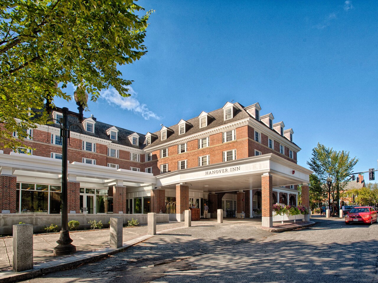 La imagen muestra un gran edificio de ladrillo, probablemente un hotel o un edificio residencial, con un letrero que dice "Hanover Inn". En primer plano hay árboles y una calle.