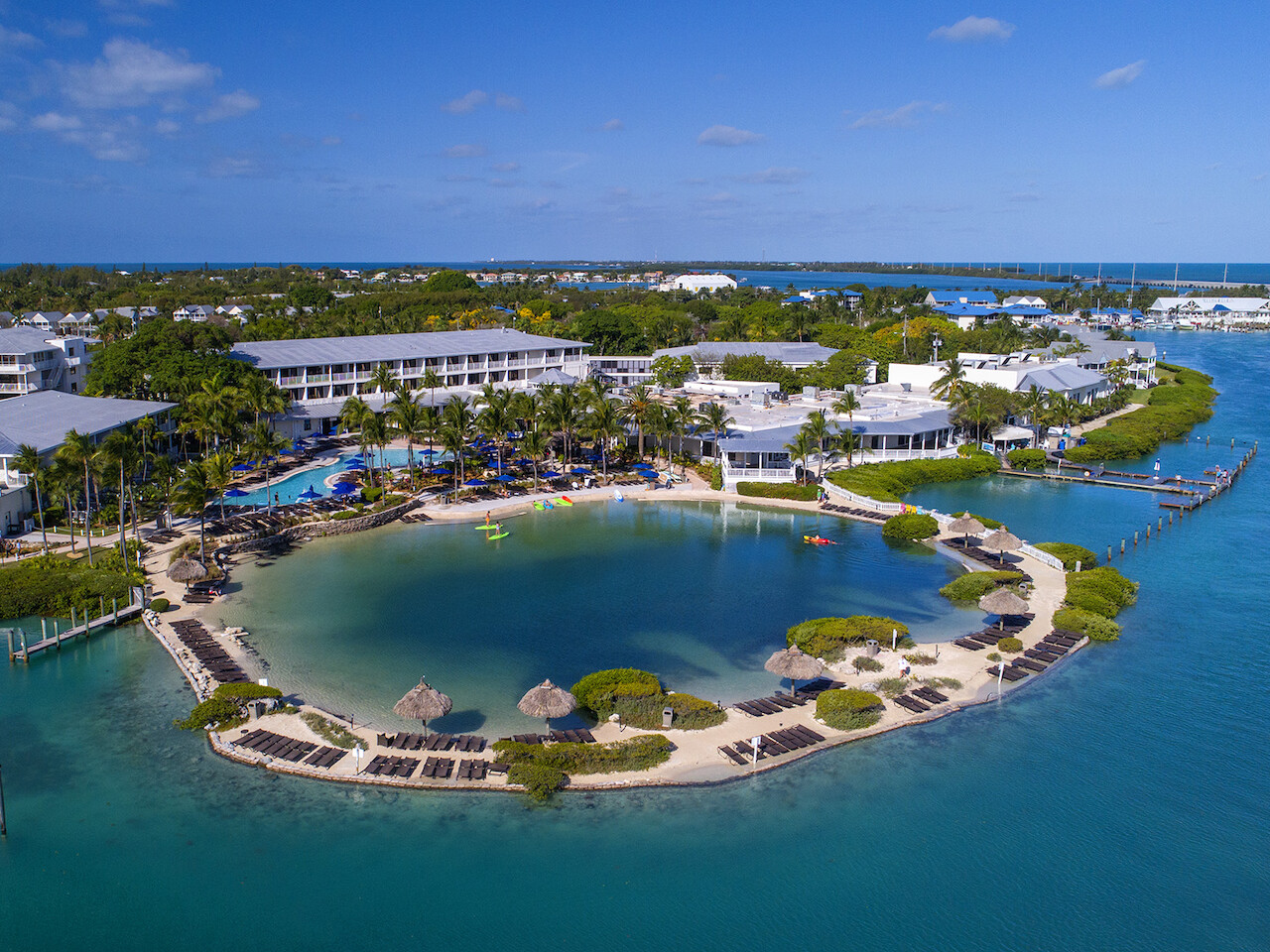 An island resort featuring lagoon-style pools, surrounding buildings, palm trees, beach chairs, and umbrellas, bordered by clear blue water and greenery.