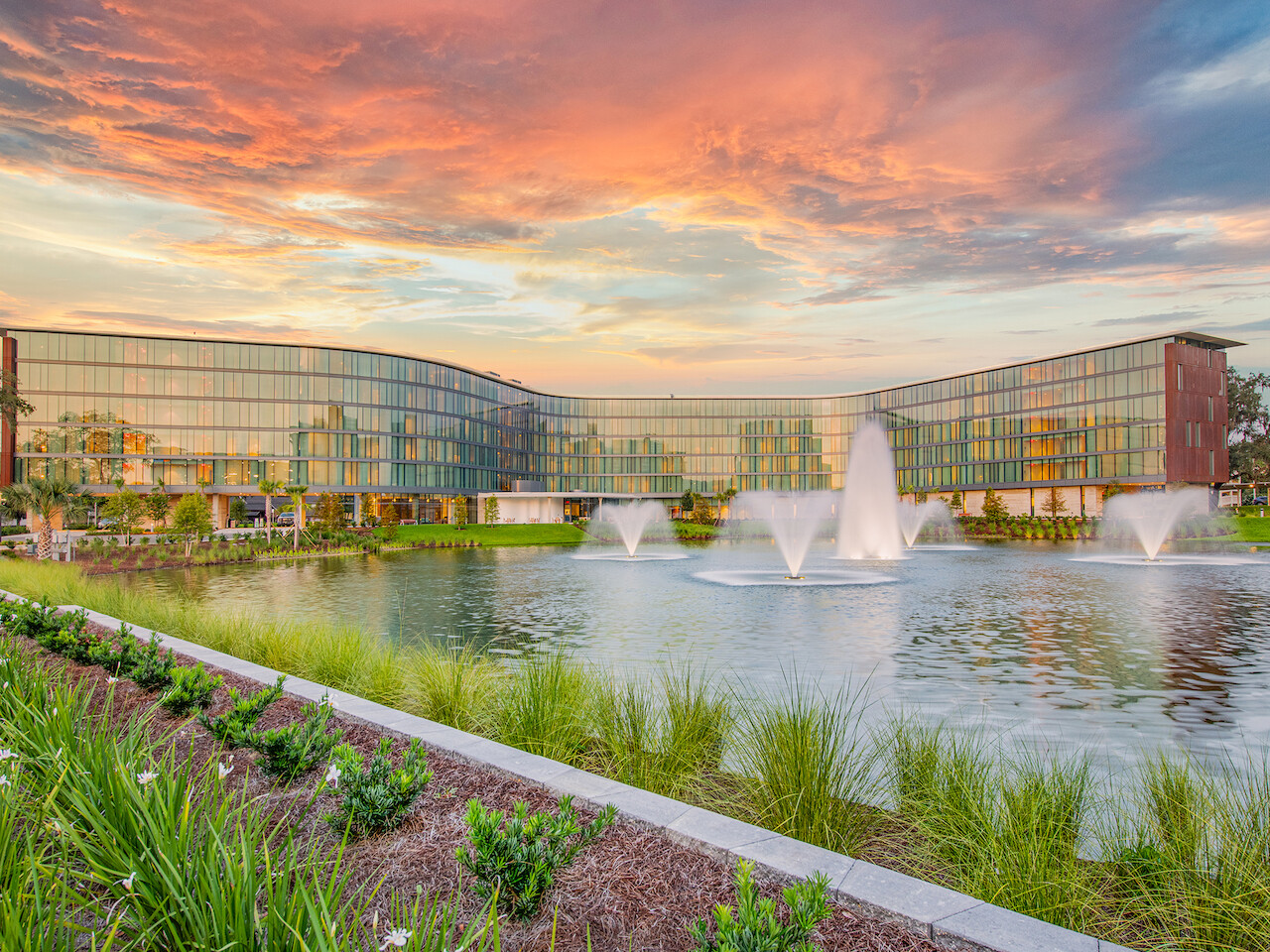 Modern building with glass façade reflected in a pond with fountains, framed by a garden under a vibrant sunset sky.