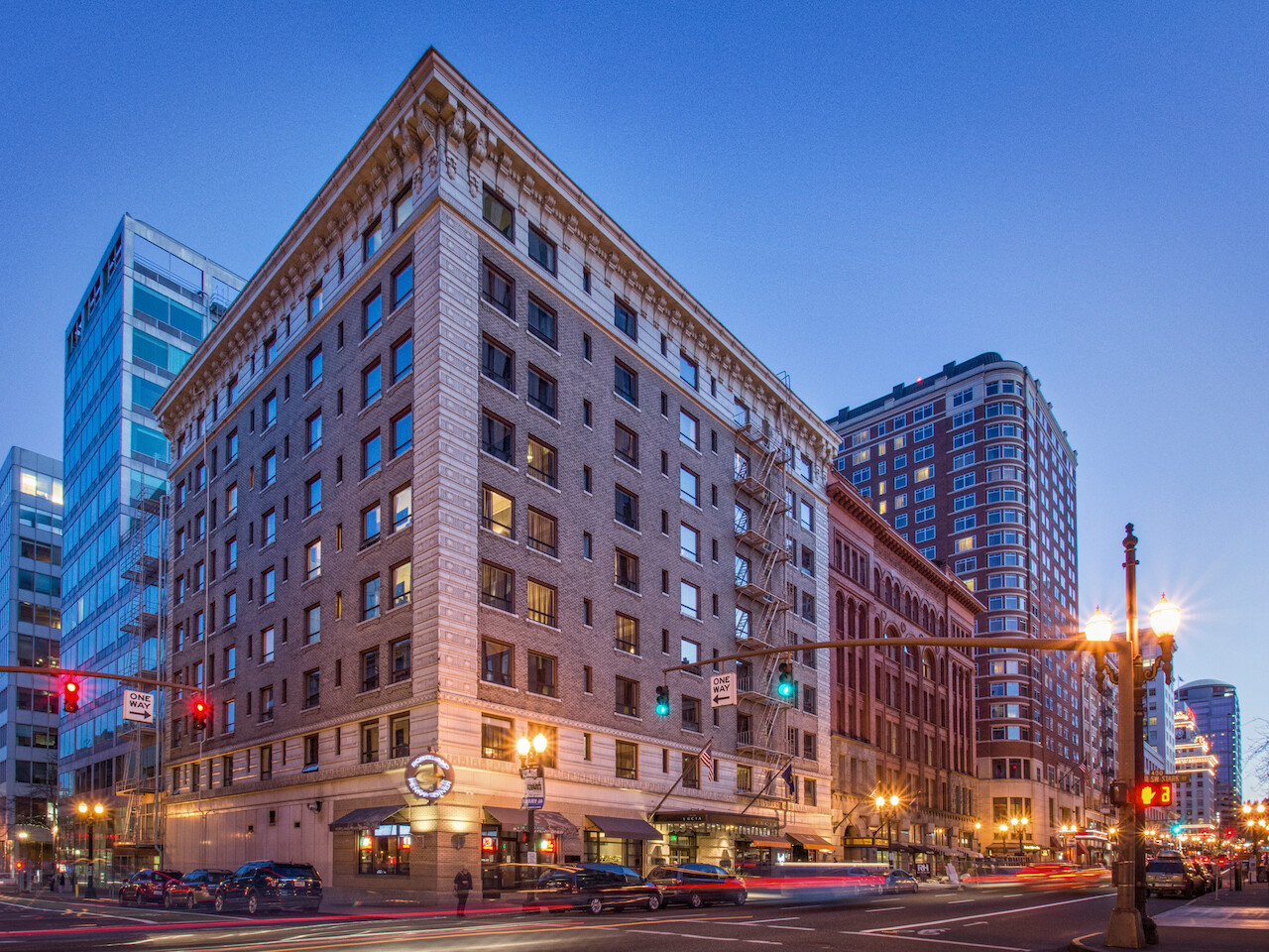 A city street scene at twilight with a historic multi-story building in the foreground, modern skyscrapers in the background, and streetlights on.