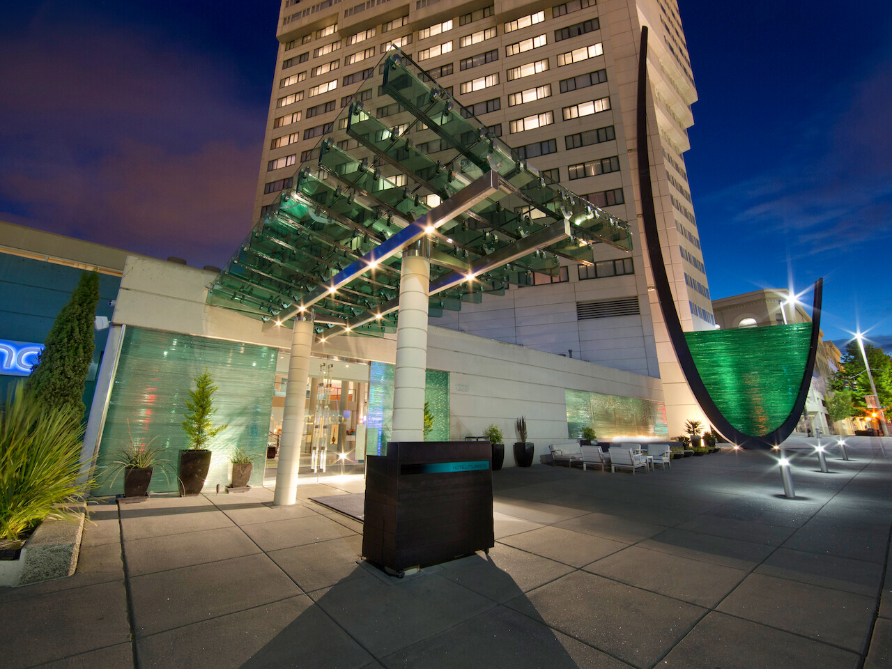 A modern building entrance is illuminated at night, featuring a glass canopy and artistic green structures, with a tall building in the background.