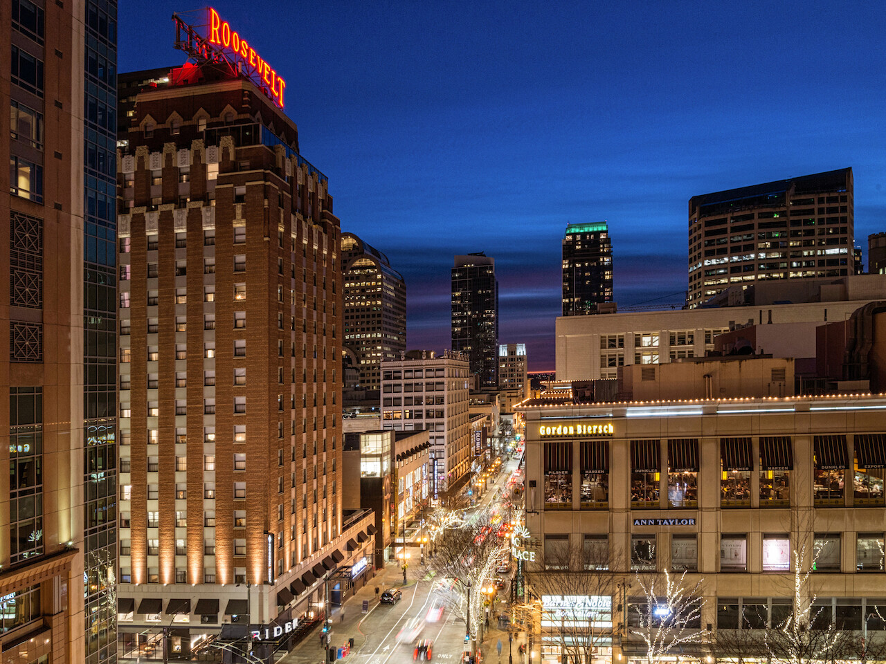 A cityscape at twilight featuring the Roosevelt Hotel and surrounding buildings, with illuminated streets and city lights.