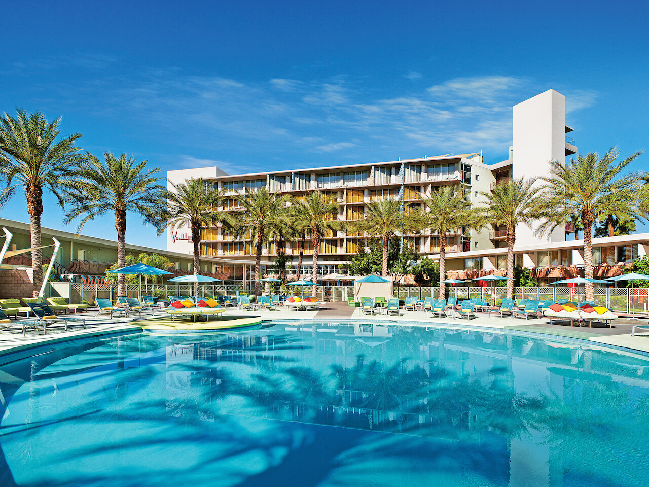 A large, inviting swimming pool is surrounded by lounge chairs, umbrellas, and tall palm trees, with a multi-story building in the background.