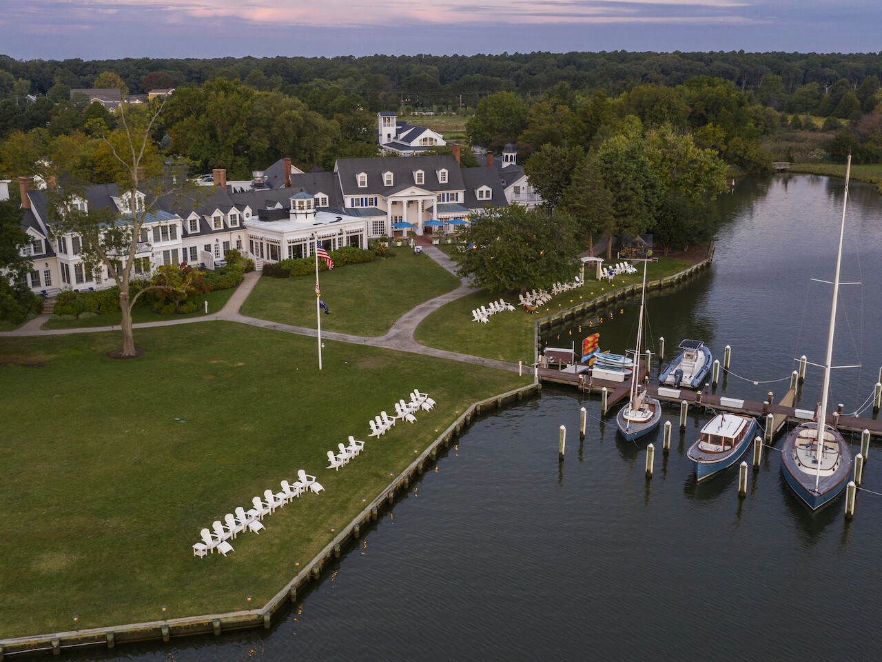 The image shows a waterfront property with a large building, a lawn with chairs, and a dock with several boats moored on the water.