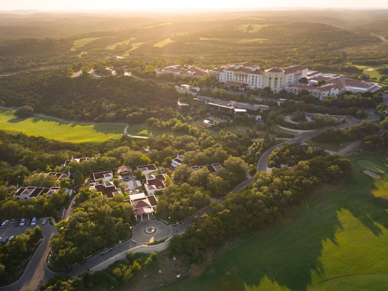 A luxurious resort with a large main building, a modern glass extension, an outdoor pool, lounge chairs, and lush greenery under a partly cloudy sky.