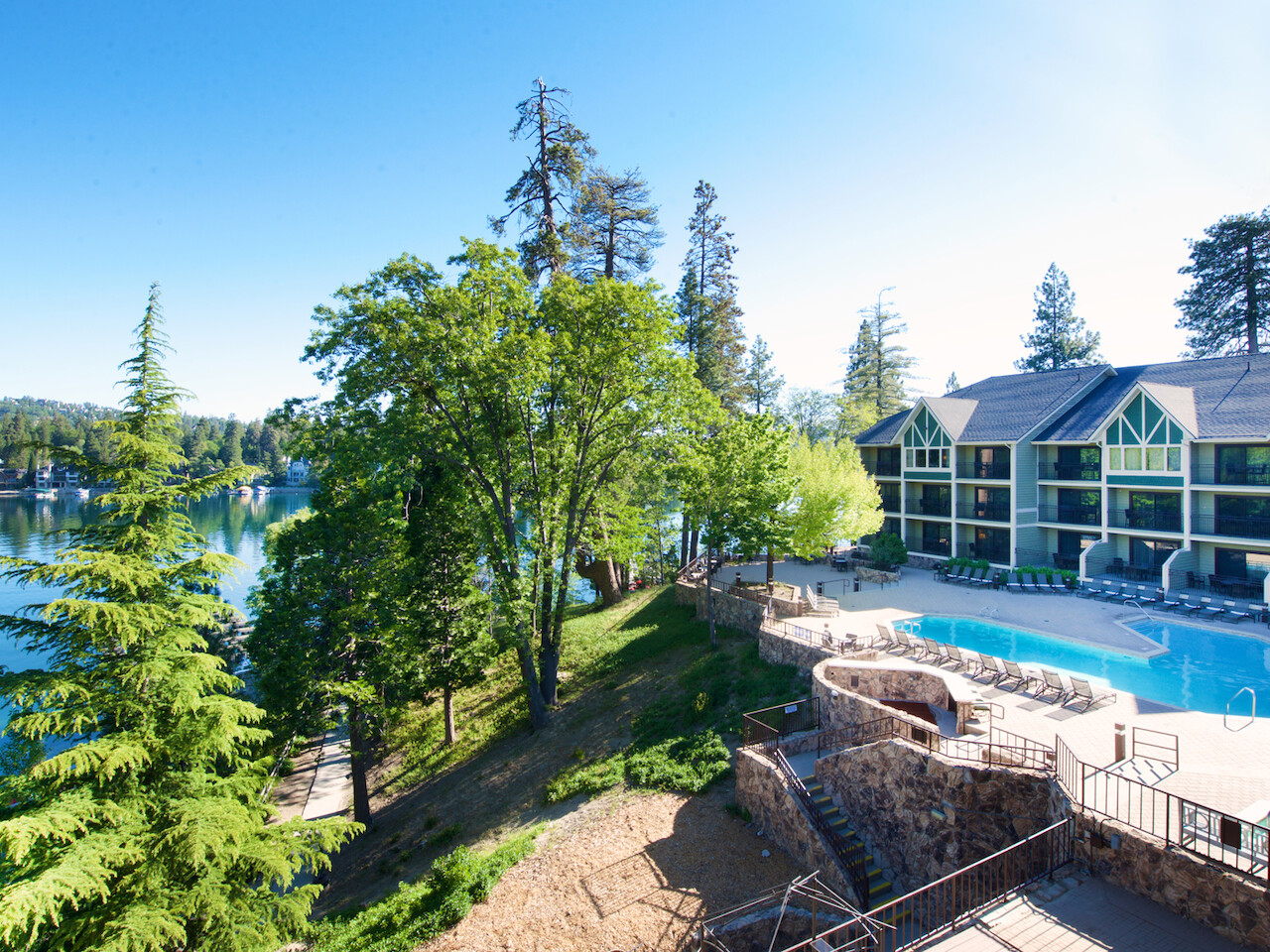 Un resort junto a un lago con árboles, edificios modernos, una piscina al aire libre y tumbonas bajo un cielo azul brillante.