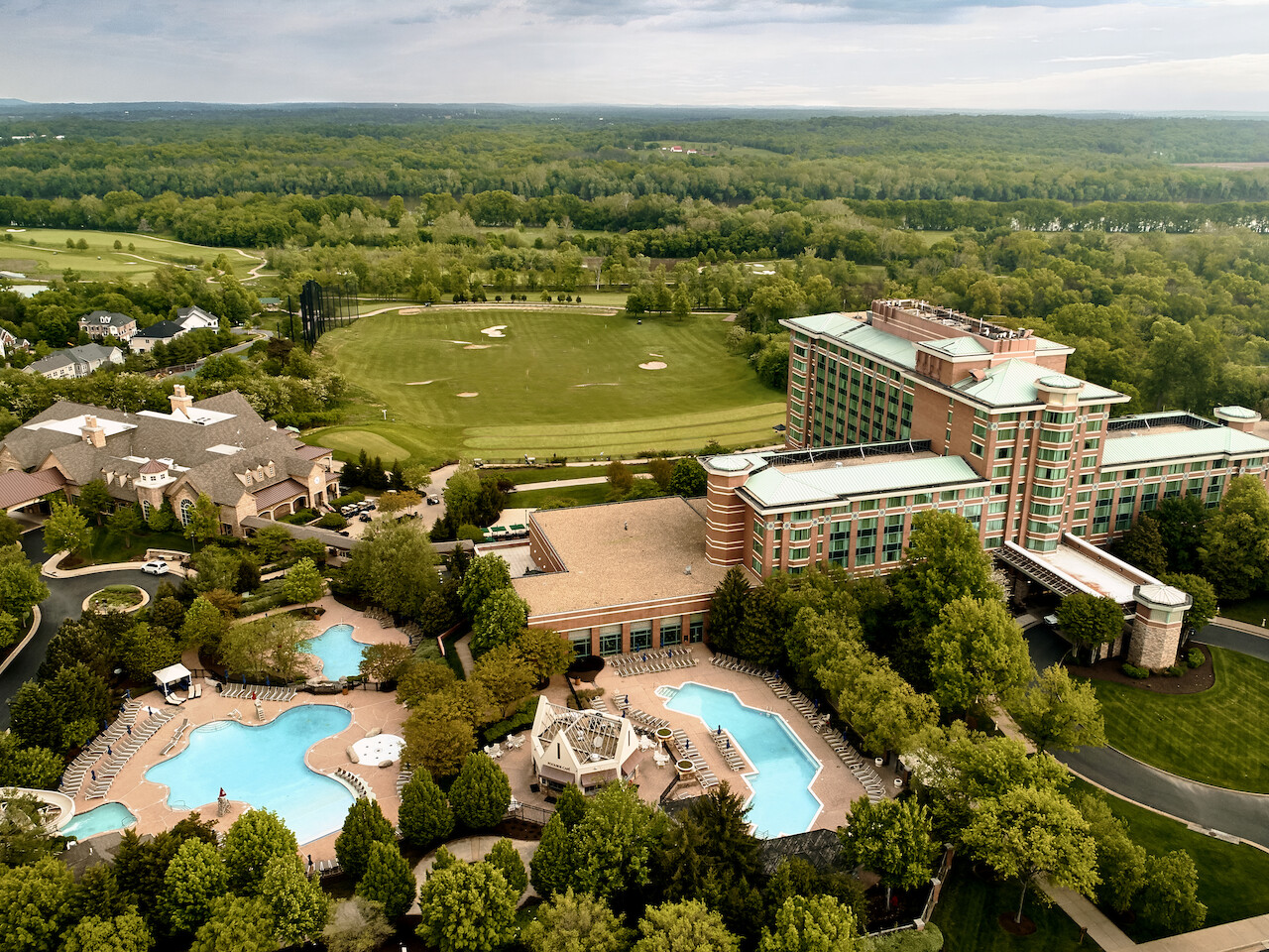 An aerial view of a large resort with multiple swimming pools, surrounded by greenery and nearby buildings, set against a vast landscape.