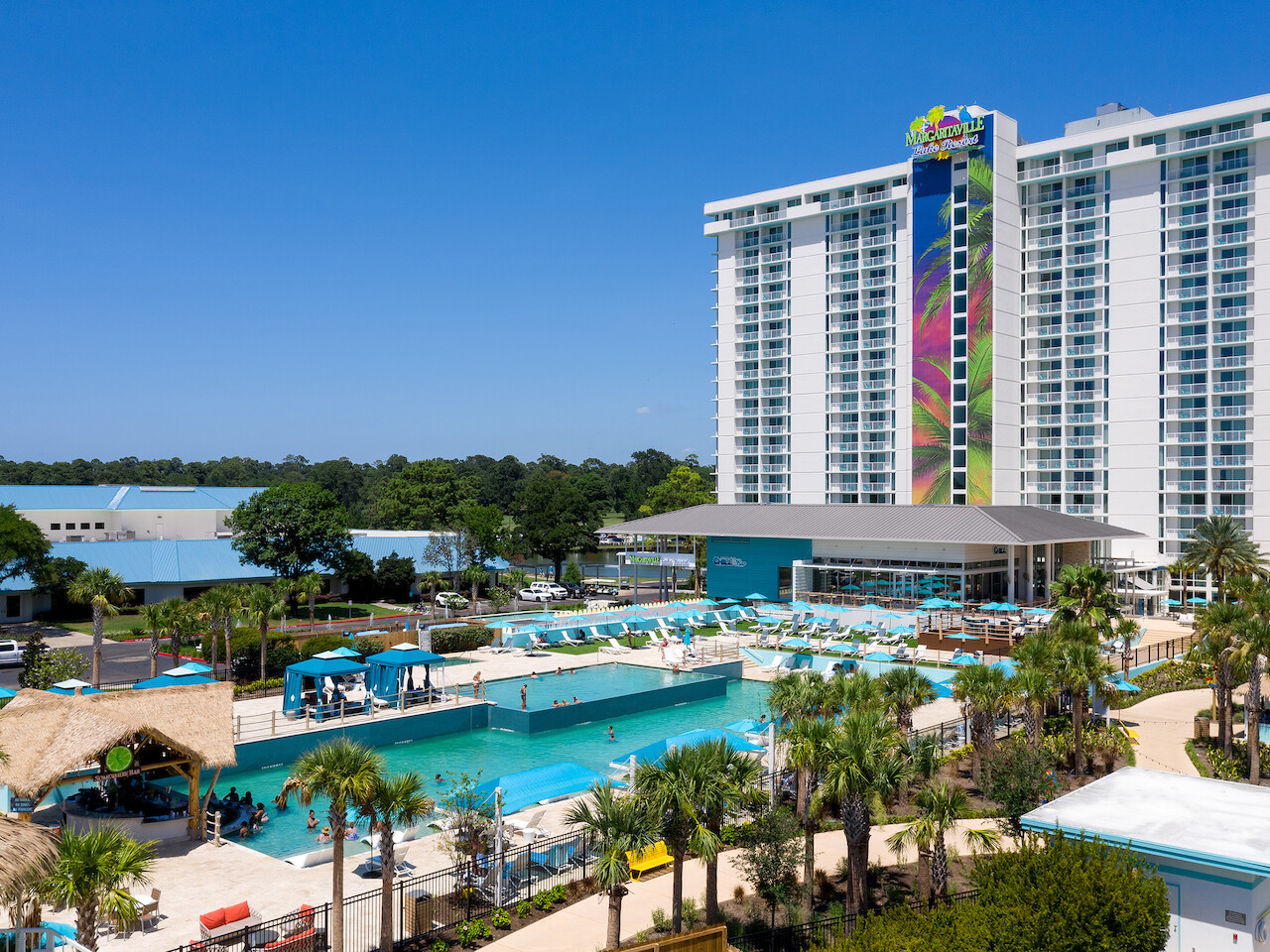 Un complejo tropical que cuenta con un hotel grande y moderno con un edificio alto, múltiples piscinas, palmeras y relajantes áreas para sentarse al aire libre.