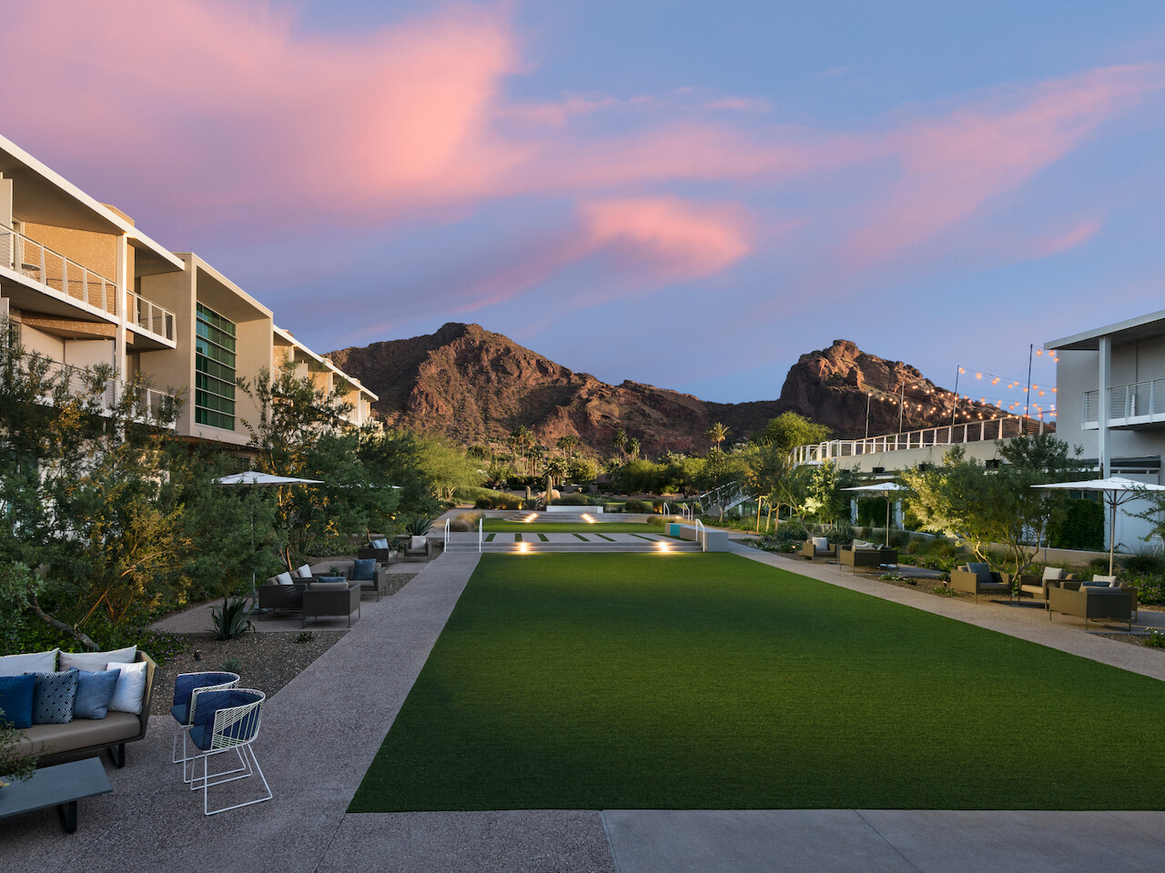 The image shows a modern courtyard with a manicured lawn, seating areas, and buildings on either side, set against a mountain backdrop during sunset.