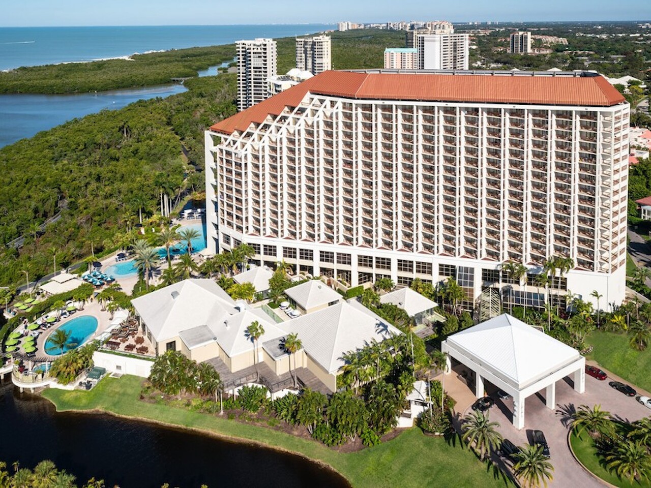 Aerial view of a large hotel with a red-tiled roof, outdoor pools, and greenery, situated near a body of water, surrounded by trees and buildings.