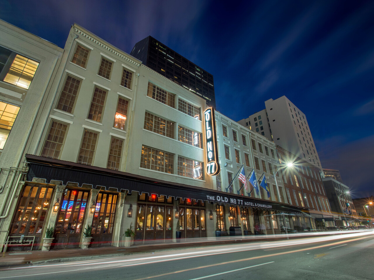 A nighttime street view featuring a historic hotel with neon signage, American flags, light trails from passing cars, and adjacent buildings.