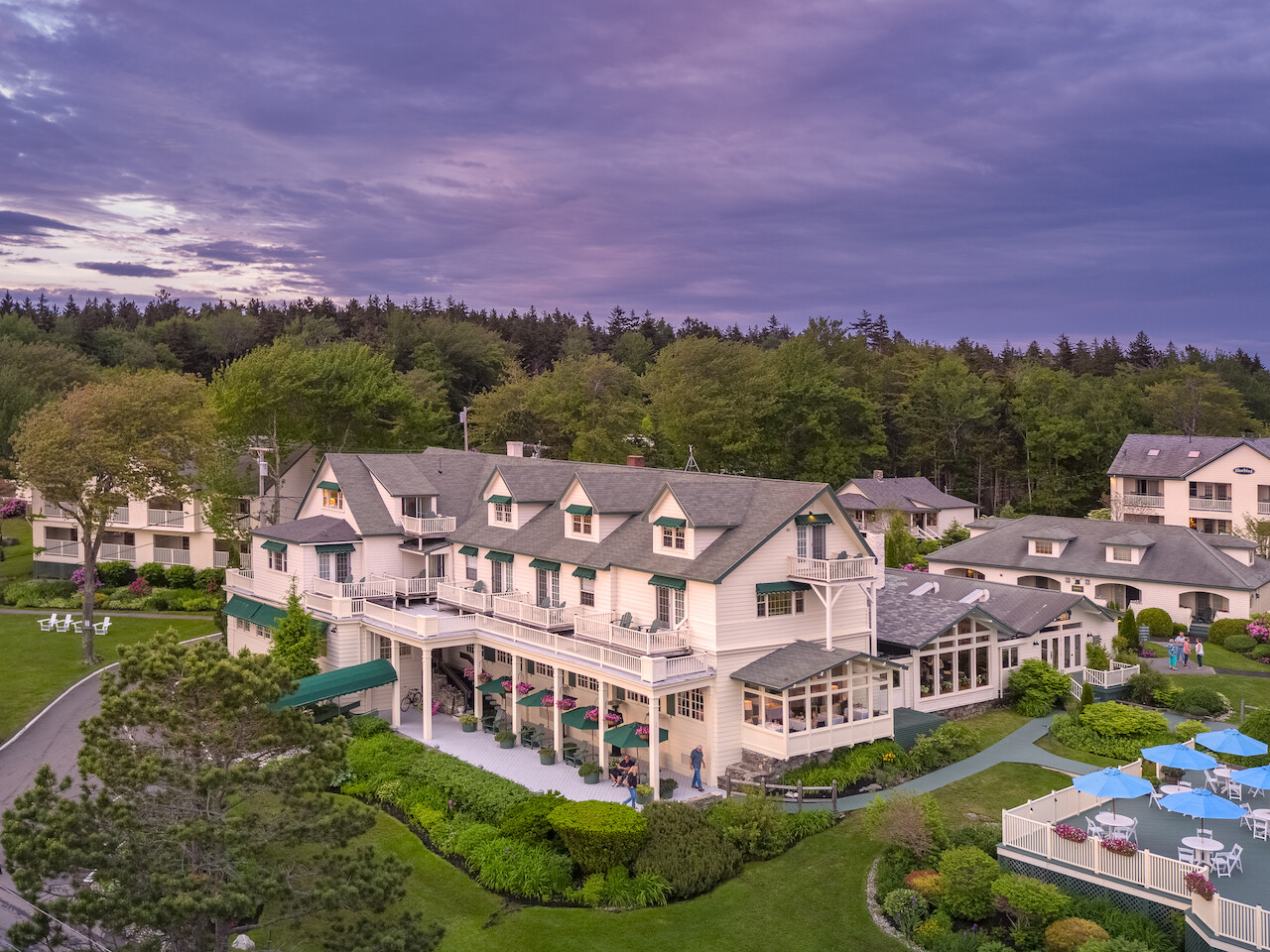 A serene hotel with classic architecture surrounded by lush greenery, featuring an outdoor pool and neatly landscaped grounds, under a purple sunset sky.