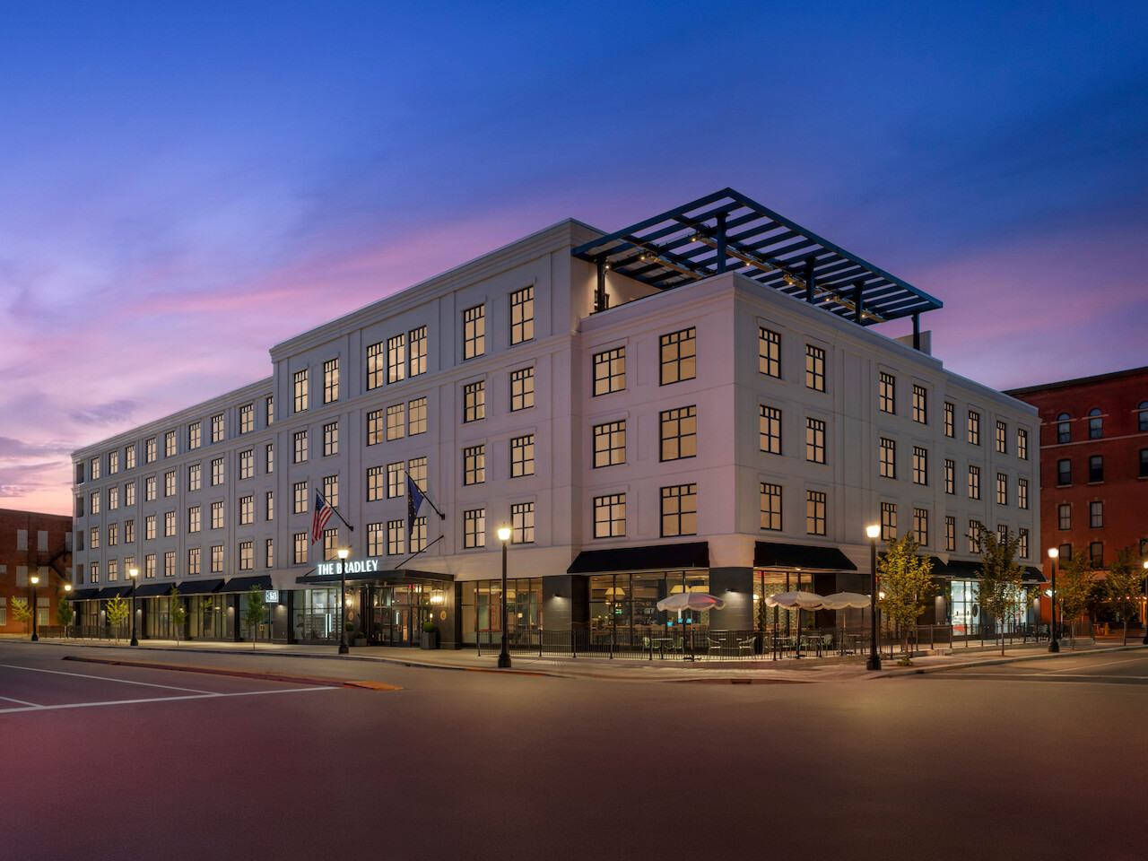 A four-story building is shown at dusk, featuring numerous windows and a rooftop structure, with lit streetlights and a few trees decorating the sidewalk.