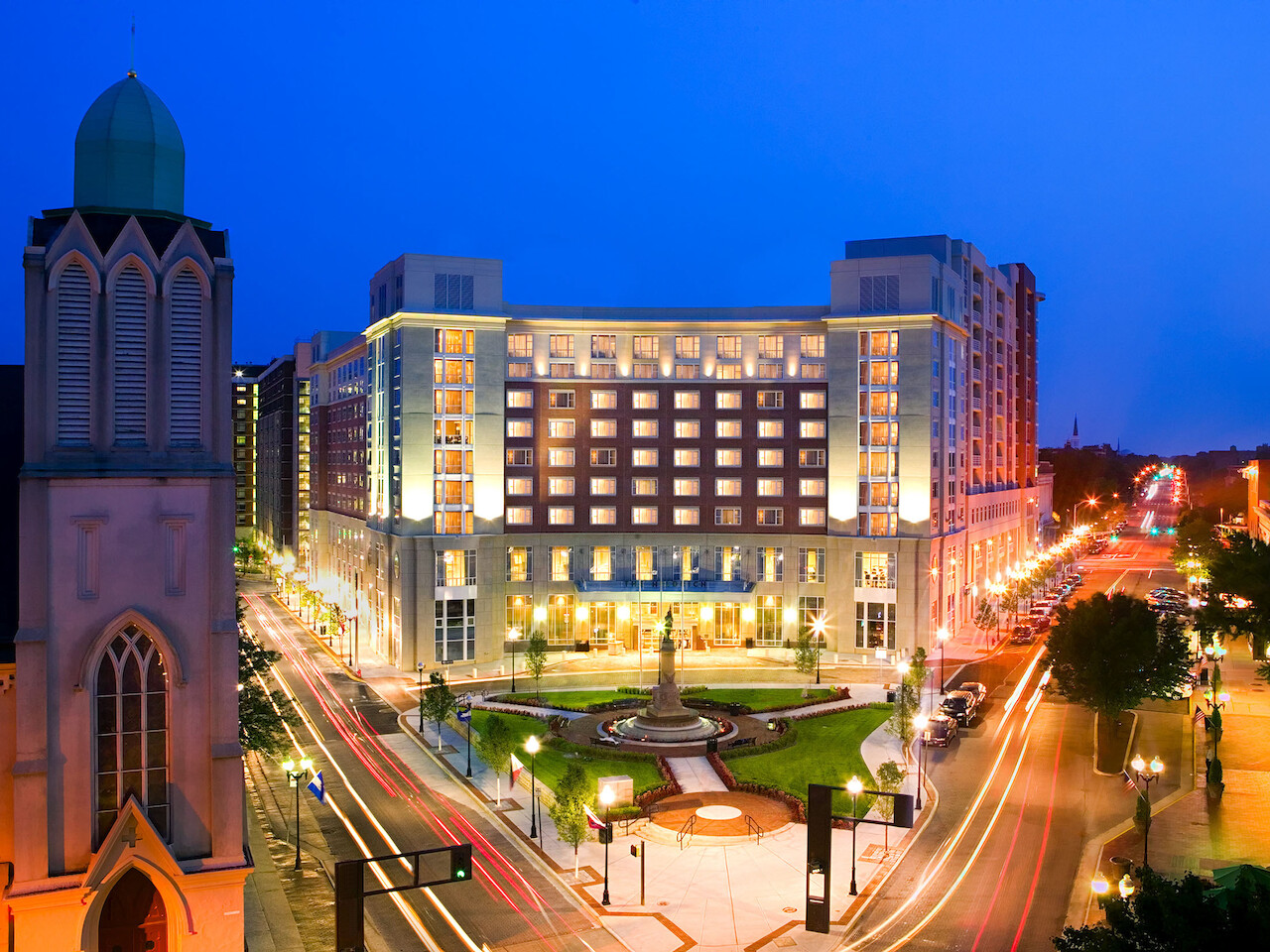 A night view of a city with illuminated streets showcases a large, well-lit building and an adjacent church, featuring a bustling urban environment.