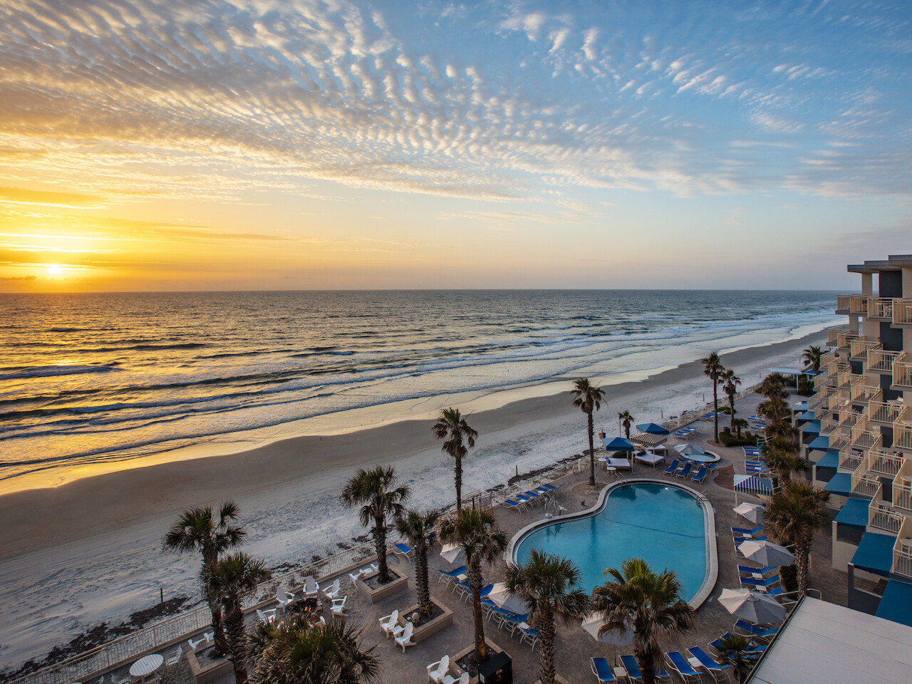 A beautiful beach scene during sunset with a pool, palm trees, and a building with balconies overlooking the ocean.