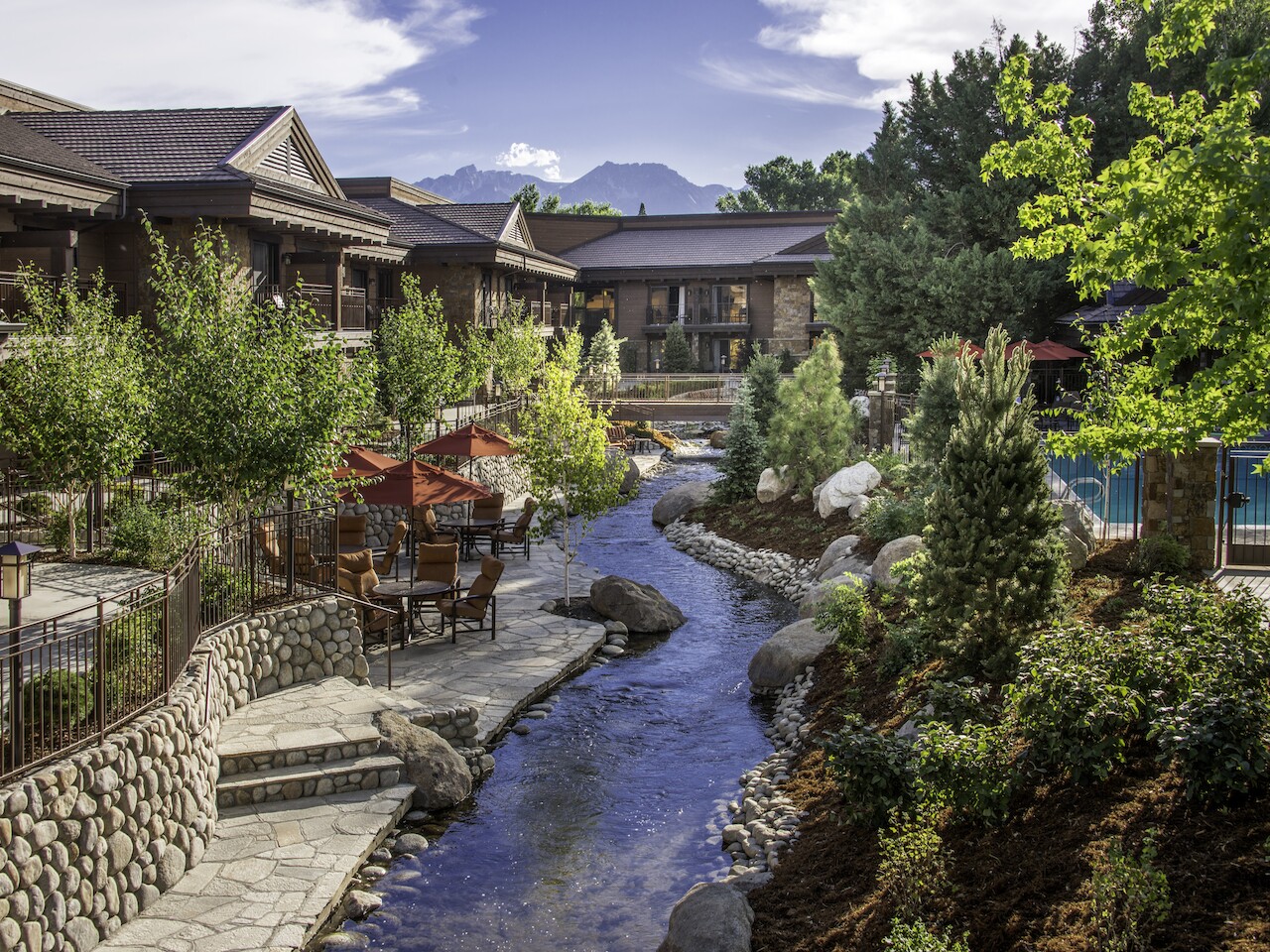 An outdoor scene with a stream running through a landscaped area, surrounded by buildings, trees, and seating areas, under a partly cloudy sky.