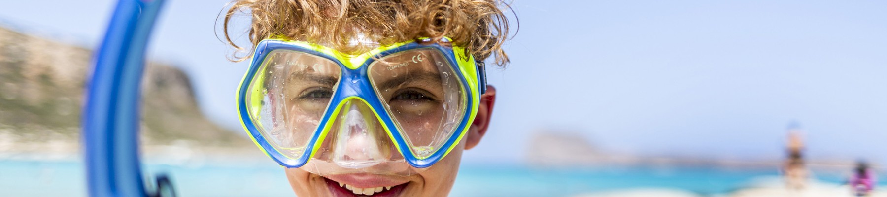 A child with curly hair is wearing a snorkel mask and holding a snorkel at the beach.