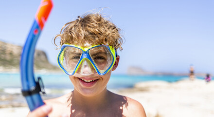 A child with curly hair is wearing a snorkel mask and holding a snorkel at the beach.