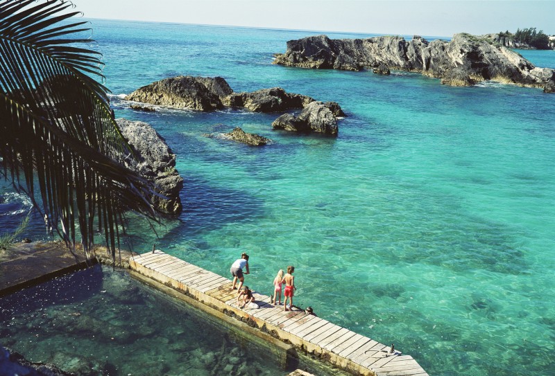A tranquil coastal scene with three people standing on a wooden dock, surrounded by clear turquoise water and rugged rocks in the distance.