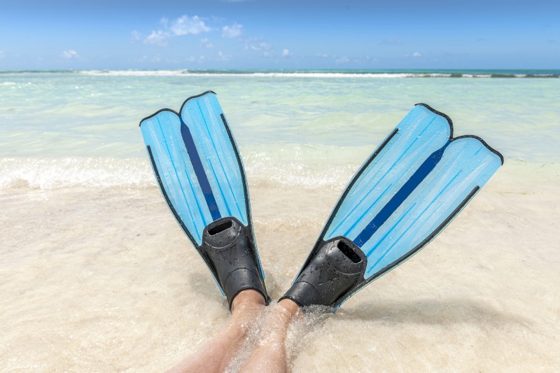 A person wearing blue snorkeling fins relaxes at the shoreline with clear ocean water and blue sky in the background.