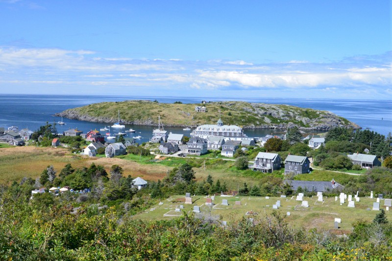 A coastal landscape featuring houses, a small harbor with boats, an island, and a cemetery in the foreground.