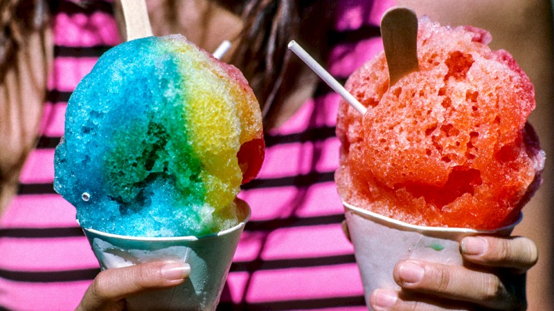 Two hands holding colorful shaved ice treats in paper cups with wooden spoons sticking out, with a person in a striped pink and black shirt.