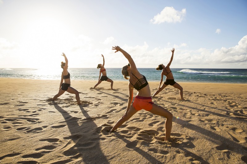Four people are practicing yoga on a sandy beach with the ocean in the background under a bright, sunny sky.