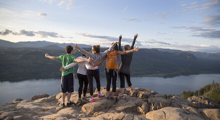 A group of people standing on a rocky cliff with arms raised, overlooking a scenic landscape of mountains and a body of water under a clear sky.