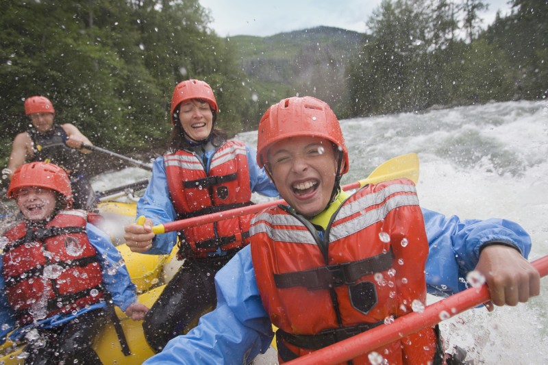 People wearing life jackets and helmets are rafting and enjoying splashes of water in a fast-moving river against a backdrop of lush green trees.
