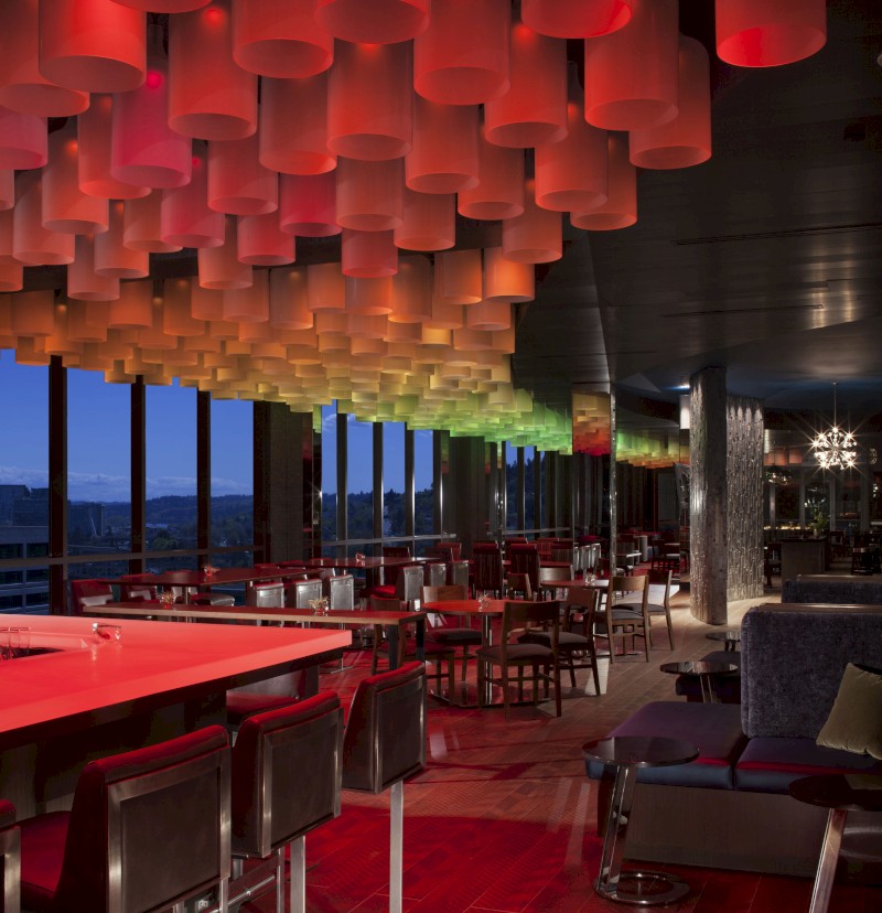 The image shows a modern, stylish restaurant interior with colorful cylindrical lights on the ceiling, large windows, and varied seating arrangements.