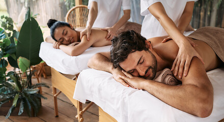Un hombre y una mujer están tumbados en camillas de masaje, recibiendo masajes en la espalda en un spa con plantas a su alrededor.