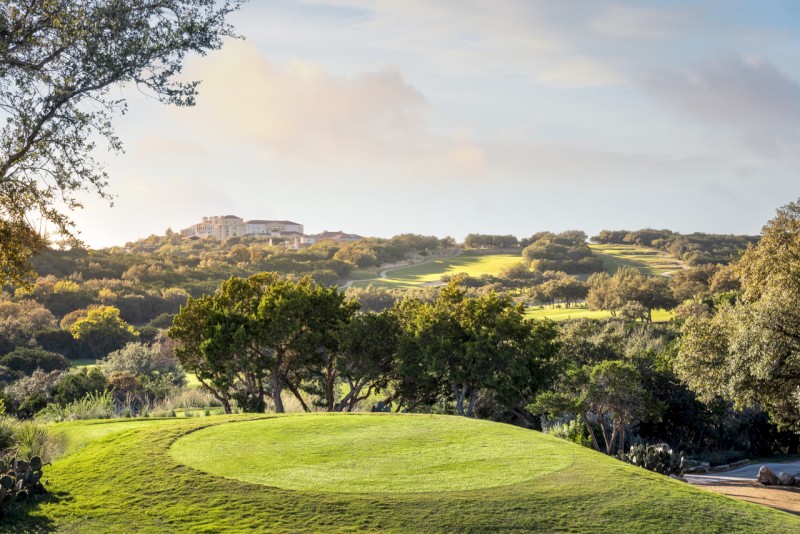 Un campo de golf con un green bien cuidado, rodeado de árboles y colinas, con un gran edificio a lo lejos bajo un cielo parcialmente nublado.