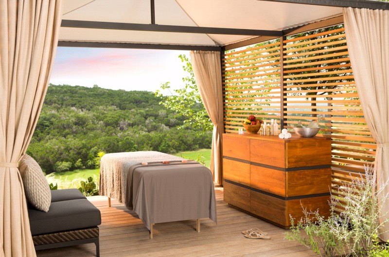 Una habitación tranquila al aire libre con camilla de masaje, cómoda y vista a frondosos árboles y vegetación, creando un ambiente sereno y relajante.