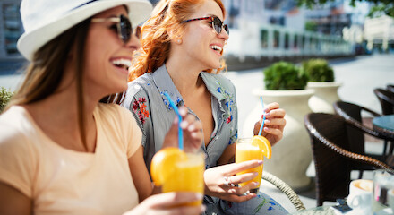 Two women wearing sunglasses are sitting outdoors, smiling, and enjoying drinks with straws, likely at a café or restaurant.