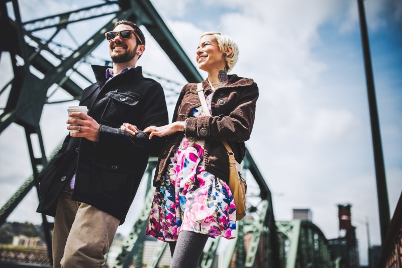 Una pareja camina del brazo sobre un puente, ambos sonriendo y disfrutando de su día. El hombre sostiene una taza de café y el cielo está parcialmente nublado.