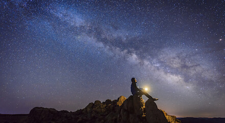 A person sits on a rocky peak under a starry sky, holding a light and gazing at the Milky Way galaxy in a dark, clear night landscape.