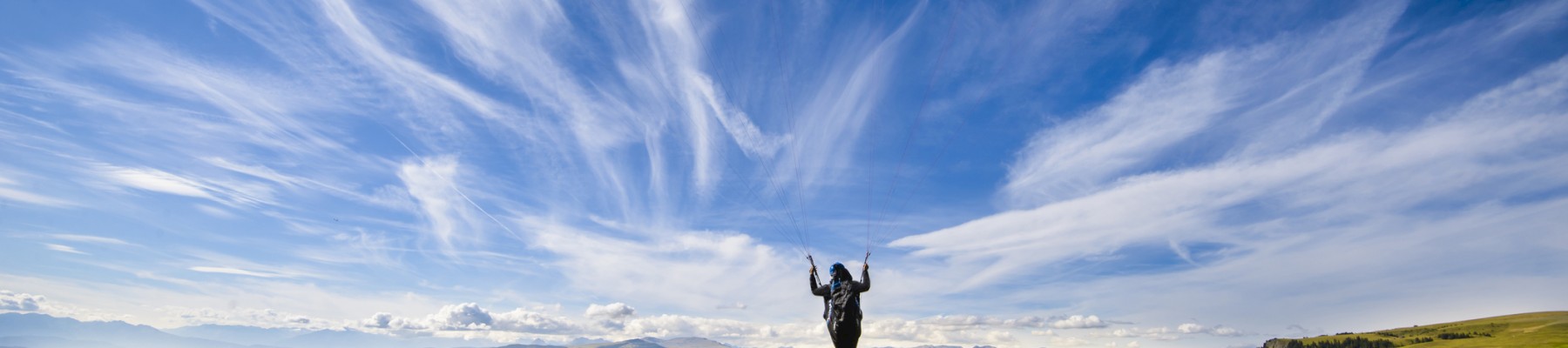 Una persona vuela en parapente sobre un paisaje vasto y exuberante con montañas, bosques y pueblos bajo un cielo azul vibrante con nubes tenues.