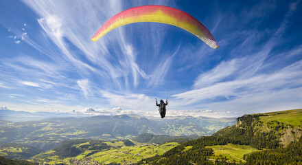 A person is paragliding over a vast, lush landscape with mountains, forests, and villages under a vibrant blue sky with wispy clouds.